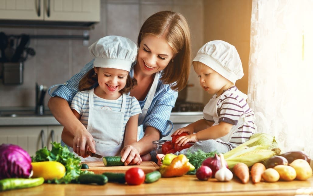 A mother and two children are in a kitchen, preparing vegetables together and smiling.