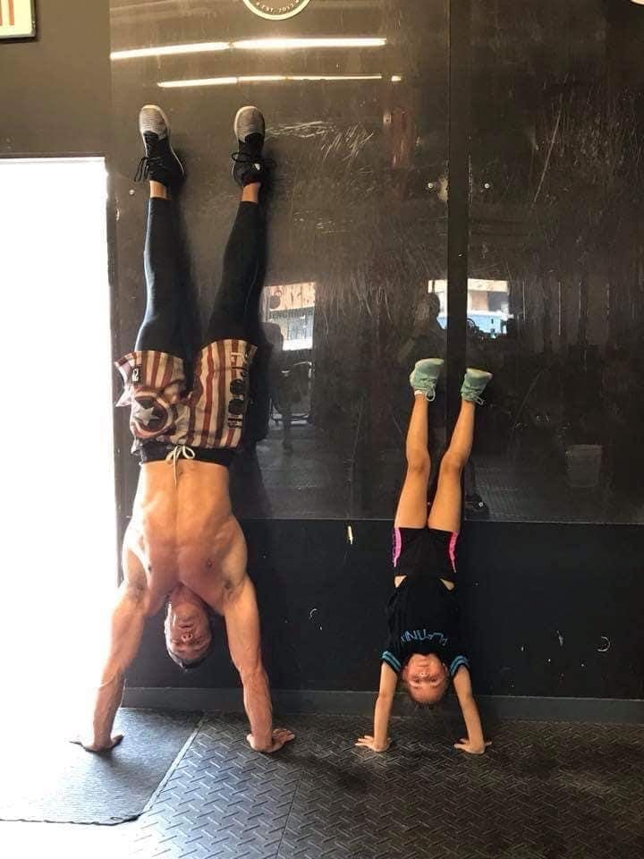 Two people are performing handstands against a wall in a gym setting.