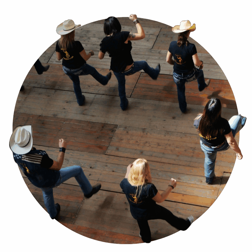 A group of people line dancing on a wooden floor, some wearing cowboy hats.